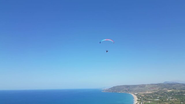 An epic panoramic drone shot of a paraglider flying under a bright blue wing over the coast of Crete. Rugged mountains and green olive groves meet the deep indigo sea. High quality 4k footage