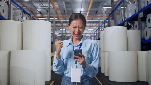 Asian Female With Her Smarphone at Warehouse with Rows of Stacked White Polymer and Paper Rolls, She Raises Her Fist Up With Screaming Goal
