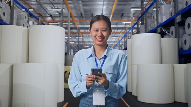 Asian Female With Her Smarphone at Warehouse with Rows of Stacked White Polymer and Paper Rolls, She Is Looking At The Camera With A Smile