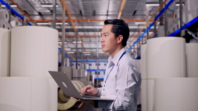 Asian Male Worker Standing at Warehouse with Rows of Stacked White Polymer and Paper Rolls, Observes By Looking Up Before He Come To Concentrating On The Laptop And Keep On Typing