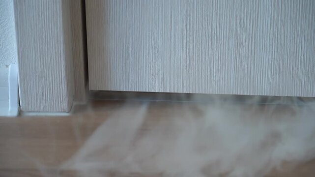 Smoke starts coming from under the door concept of fire smoke ignition. The living room has a close-up view of the entrance door, from under which thick white smoke begins to abruptly rise. High