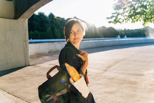 Attractive young woman with baguette in backpack on sunny city park