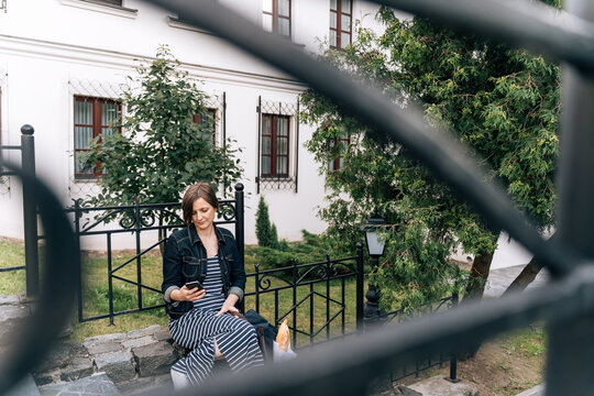 Woman on stone stairs using phone, backpack with baguette nearby