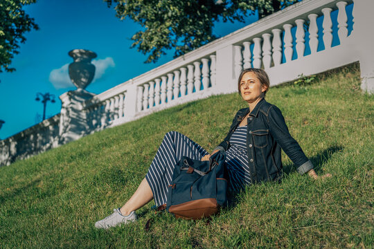Woman in striped dress and denim jacket sitting on grass with backpack