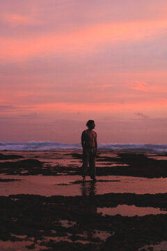 Man standing on wet rocky beach at pink and purple dusk
