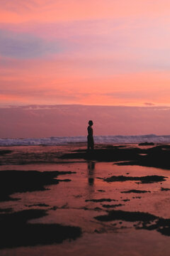 Lone man silhouette standing on rocky shore at pink and orange dusk