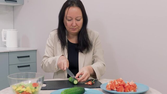 Woman in blazer chops fresh cucumber on cutting board in her kitchen. She prepares healthy vegetable salad for lunch or dinner. Lifestyle video showing cooking process and healthy eating habits