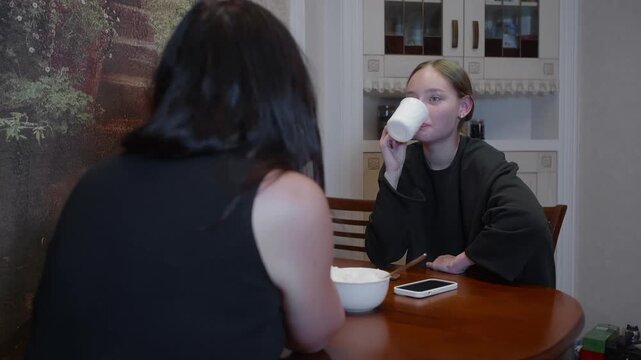 Two young women sit at dining table having breakfast. One woman shares news or shows something, causing her friend to react with shock, surprise, and excitement. Candid moment at home