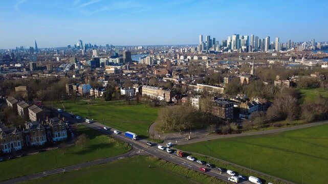 Rising London Blackheath Drone shot with view of Greenwich. Shard and Canary Wharf cityscape in the background. Blackheath in foreground with road and traffic cutting through. Mix of buildings.