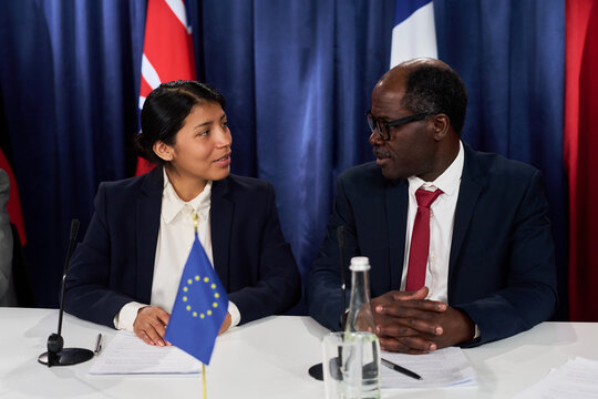 Hispanic woman and African American man discussing documents during international press conference, representing diplomacy and policy negotiation