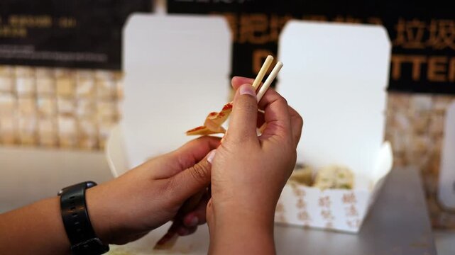 Person opens wrapper on chopsticks to eat dumplings from takeout box.
