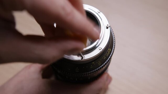 Close-up of hands unscrewing the mount on a cinema lens to swap from EF to PL bayonet. Detailed lens modification process shown on a wooden desk. Real hands-on camera gear footage.