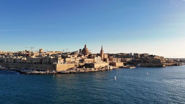 Basilica of Our Lady of Mount Carmel rises over Valletta Malta skyline with limestone buildings and Grand Harbour waterfront as camera slowly pushes in, drone gliding foward fron Mediterranean Sea