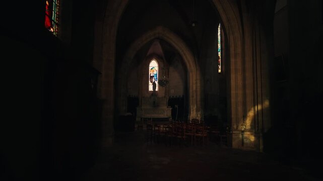 Sunlight filters through a stained glass window into the dark interior and stone arches of a historic church in Issigeac, Dordogne. Low angle view.