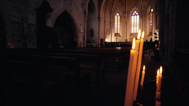 Flickering candles stand in the foreground of a quiet church interior in Monpazier, Dordogne, with wooden pews in the distance. Slow motion traveling shot.