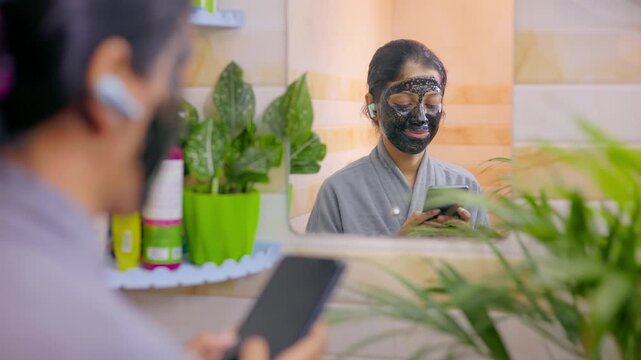 young woman relaxing with charcoal face mask, enjoying music through Bluetooth earbuds and holding mobile phone.