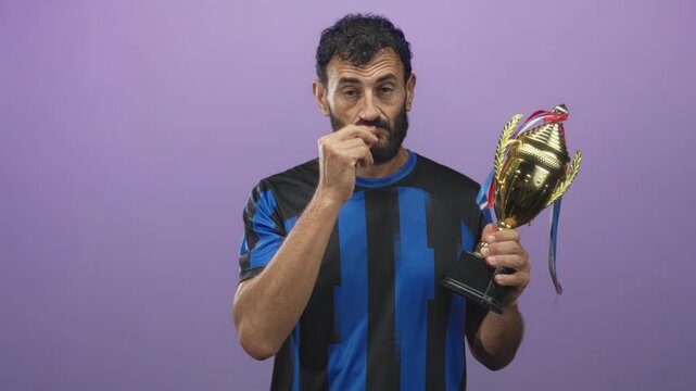 Man in soccer uniform holds gold trophy and touches mouth with hand in studio; contemplation modest pride.