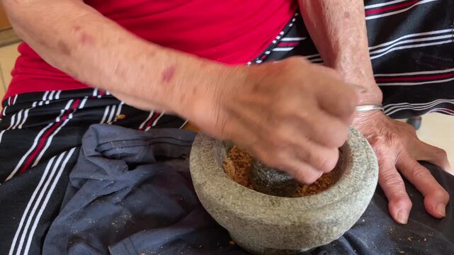 A grandmother's hands using a stone pestle to rhythmically pound and grind roasted peanuts inside a mortar, capturing a traditional manual technique for handmade food.