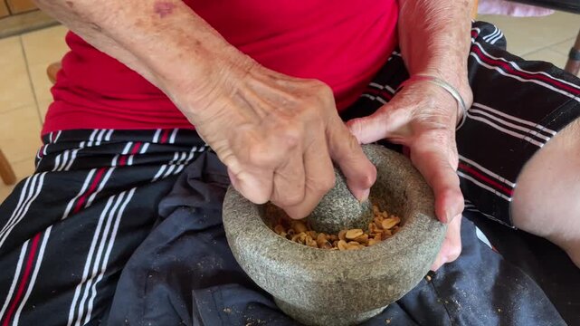 A senior woman's hands using a stone pestle to crush, grind, and pound roasted peanuts into coarse pieces within a traditional mortar.