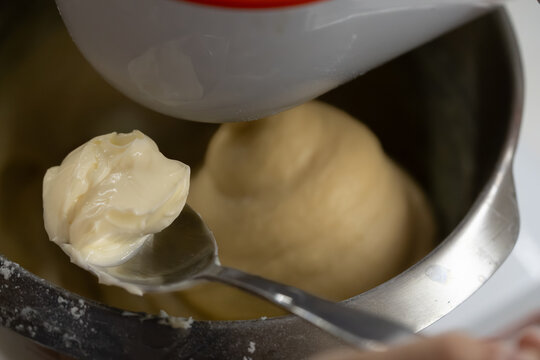 Series of photos showing the process of adding soft butter into dough. Dough in a planetary mixer metal bowl. A tablespoon with butter held above the bowl. Home cooking. 