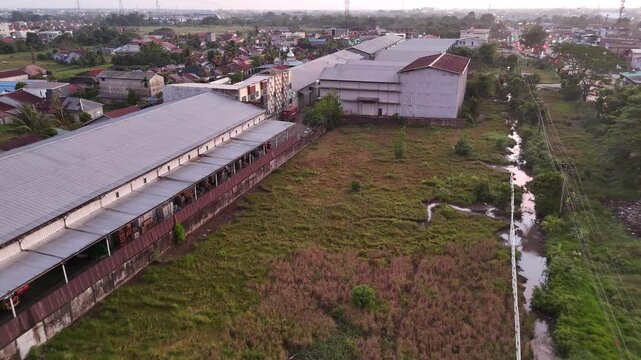 A drone slowly pulls back, revealing large metal-roofed warehouses, drainage channels, green fields, the densely populated neighborhoods surrounding them, and a city in the distance.