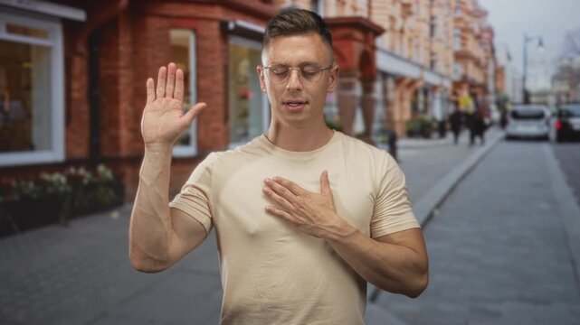 Man raises right hand and then places left hand over chest while wearing glasses on a city street; pledge sincerity.