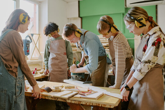 Wide shot of diverse teenagers learning woodworking, operating power tool under instructor guidance in workshop. Useful for vocational education, STEM training, skilled trades promotion