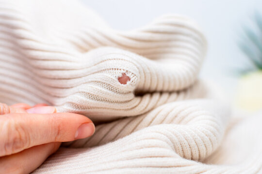 Clothing repair, craft sewing and textile recycle. Woman hands holding light beige knitted texture cloth with hole made by moth or ripped, close-up. Tailor holding damaged fabric with loose loops.
