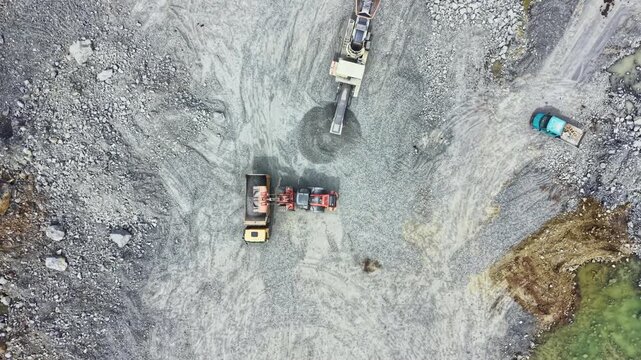 Workers are using heavy machines to extract granite from a quarry. Trucks are collecting the stone and a conveyor belt moves the material in the area.