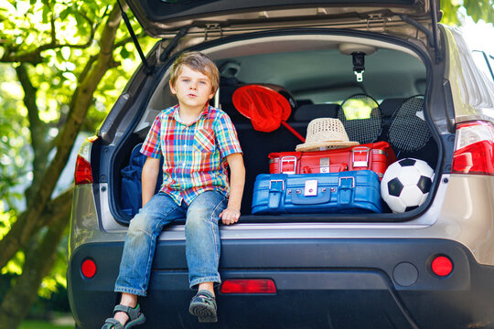Adorable little kid boy sitting in car trunk just before leaving for summer vacation with his parents. Happy child with suitcases and toys going on journey, real life, real people