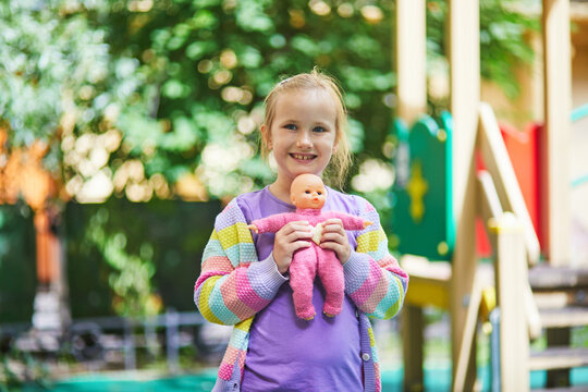 Happy young girl standing on a playground and holding a baby doll, capturing childhood, play, imagination and outdoor leisure in a colorful park setting