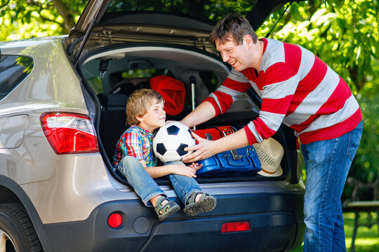 Adorable little boy sitting in car trunk as dad packs suitcases. Happy family preparing for summer vacation, cheerful children, real life, travel adventure