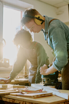 Vertical shot of young adult Caucasian man operating power router in woodworking class while Black woman assisting nearby. Supporting vocational training, skilled trades education, vertical