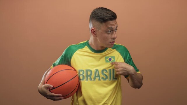 Man holds basketball with left hand and points finger to brazil flag patch on yellow jersey in studio while looking to the side across frames; pride.