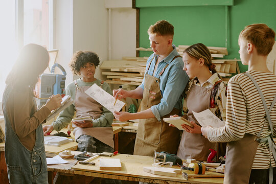Multiethnic teenagers reviewing woodworking plans and taking notes during carpentry class, developing practical skills and teamwork for vocational education marketing and training