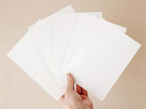 Hand holding fan of blank white vertical cards mockup on neutral beige background. Minimalist stationery or invitation suite template