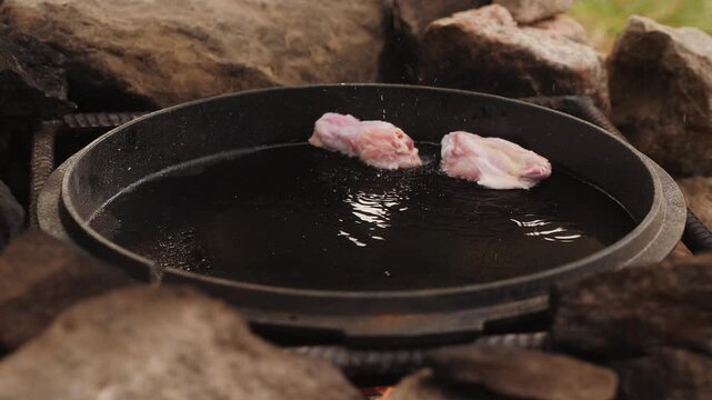 Chef placing raw chicken wings into a hot frying pan with oil, cooking meal on open fire outdoors among rocks.