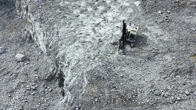 Heavy machinery works in a granite quarry, removing large amounts of stone from the ground. The area shows signs of excavation, with rocks and earth surrounding the equipment.