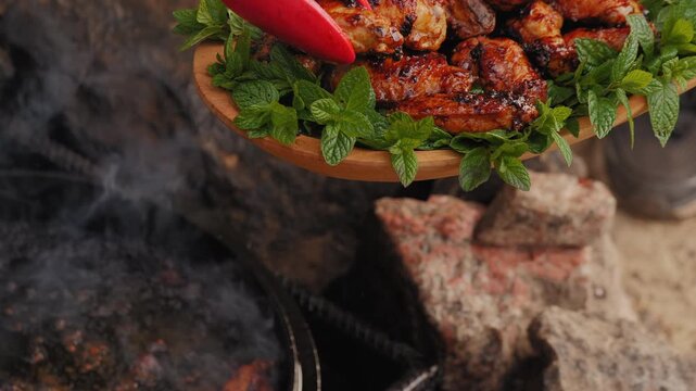 Roasted BBQ chicken wings on a wooden platter with fresh mint leaves, chef taking meat off the fire with tongs.