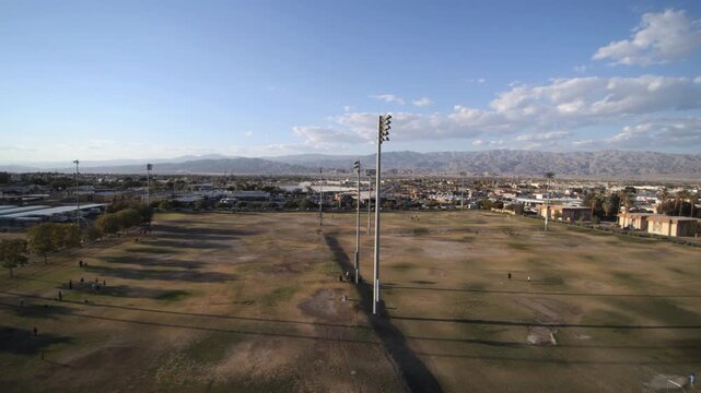 Aerial drone shot of vast sports fields with stadium lights in Indio, CA. Mountain backdrop, blue sky, and urban sprawl visible in the distance.
