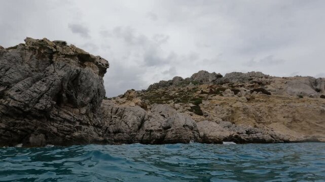Low water-level view of dramatic rocky limestone cliffs along the Souda coastline in Chania, Crete, Greece. Turquoise Mediterranean water foreground, overcast sky.
