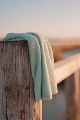 Fototapeta premium Mint green towel draped over a wooden pier post at sunset. Coastal summer lifestyle concept. Relaxing vacation mood by the ocean. Soft evening light on fabric texture and wood grain.