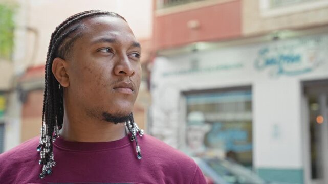 Man with braided hair and silver beads, face visible and looking to the side by a storefront on a street; quiet contemplation.