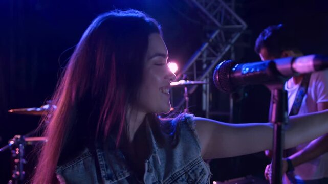 Singing vocalist wearing denim vest reaching mic stand onstage, confetti falling, guitarist playing