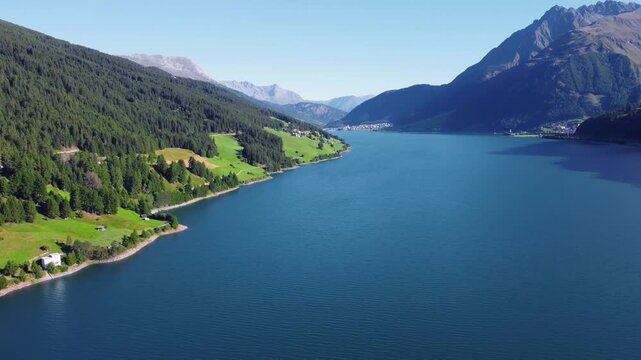 Aerial panorama view of Lake Resia and the lush Val Venosta valley in the Italian Alps. Scenic summer landscape with blue water, green fields, and majestic mountains under a clear sky