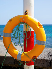 Fototapeta premium Orange lifebuoy attached to pole by the sea with rope and clear blue water in coastal safety setting