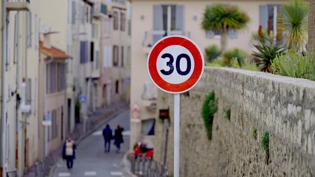 A round speed limit 30 sign on a white pole, with people walking along a narrow street in motion.