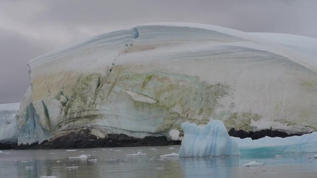 Cinematic moves slowly left past a massive ice cliff stained with orange and green penguin guano at Georges Point, Rong&eacute; Island, Antarctica