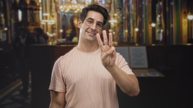 Man with glasses smiling, wearing pink shirt, showing three fingers and presenting palm toward ornate bar counter in building; friendly guidance.