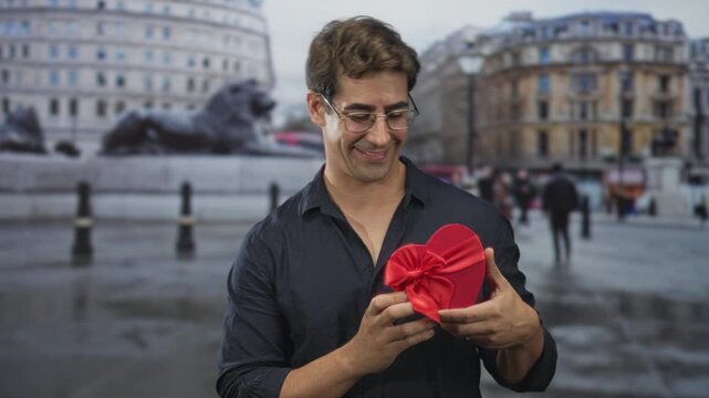 Man holds red heart shaped gift box with both hands while smiling and looking down in a busy city street with stone lion statues and historic buildings; romantic love.
